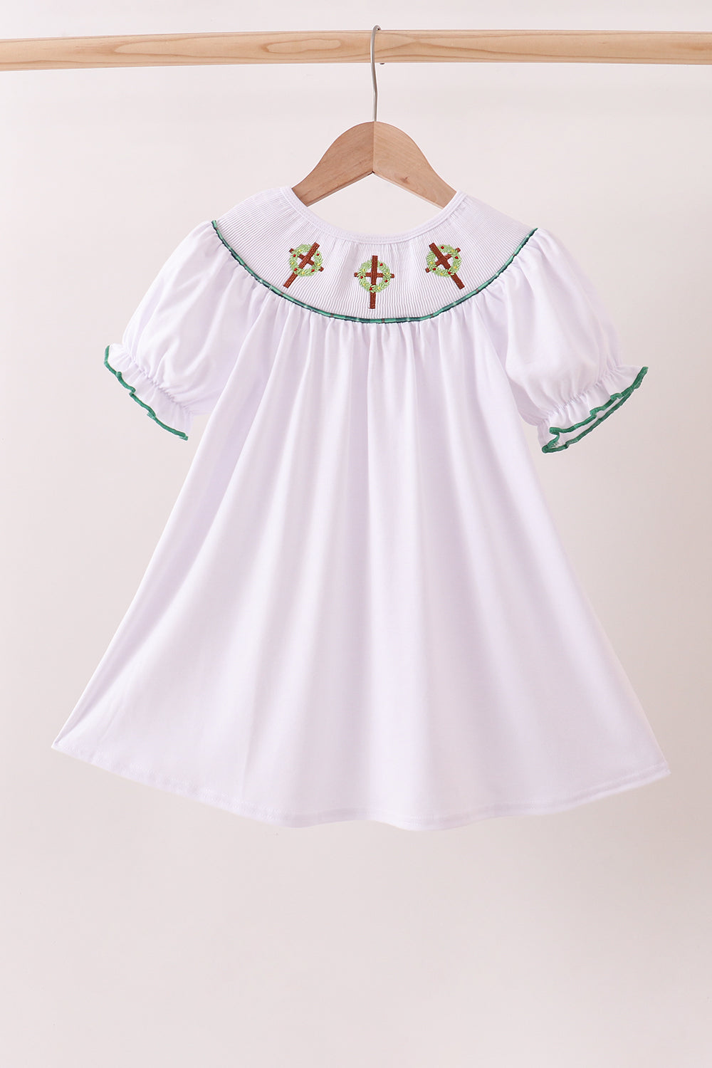White dress with green trim and red embroidery on a hanger against a beige background